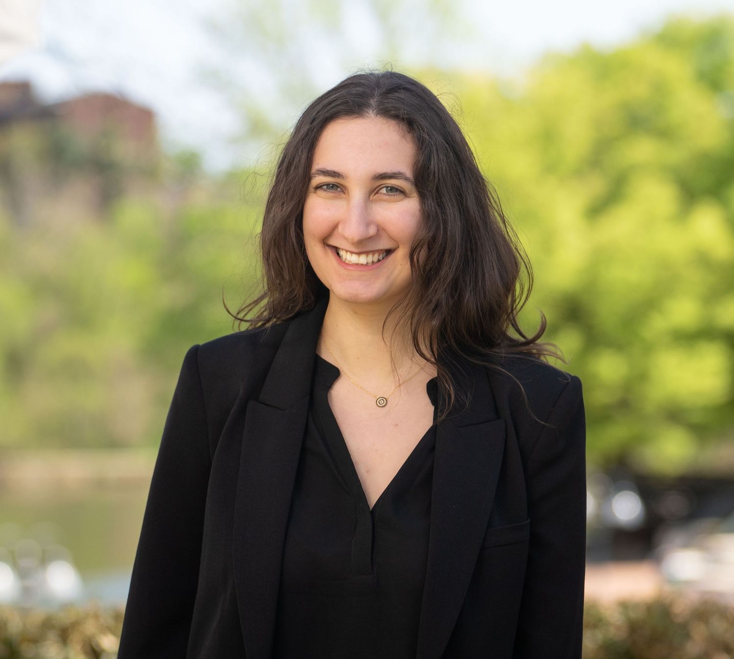 Professional portrait of someone in black business attire with a natural outdoor background.