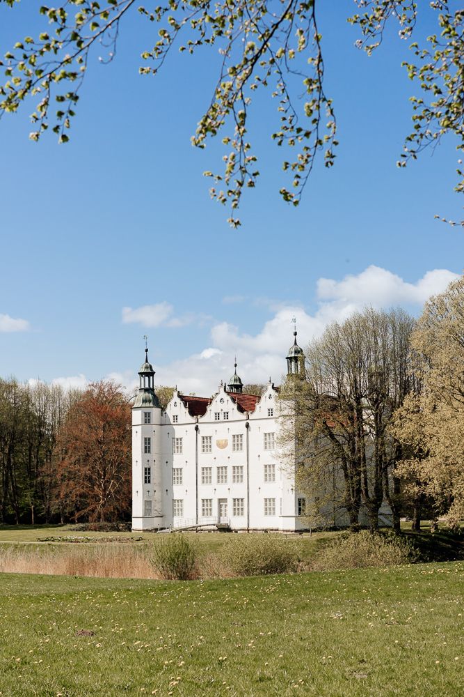 A panoramic view of a historic white manor house with Tudor-style architecture set against a spring meadow and blue sky.
