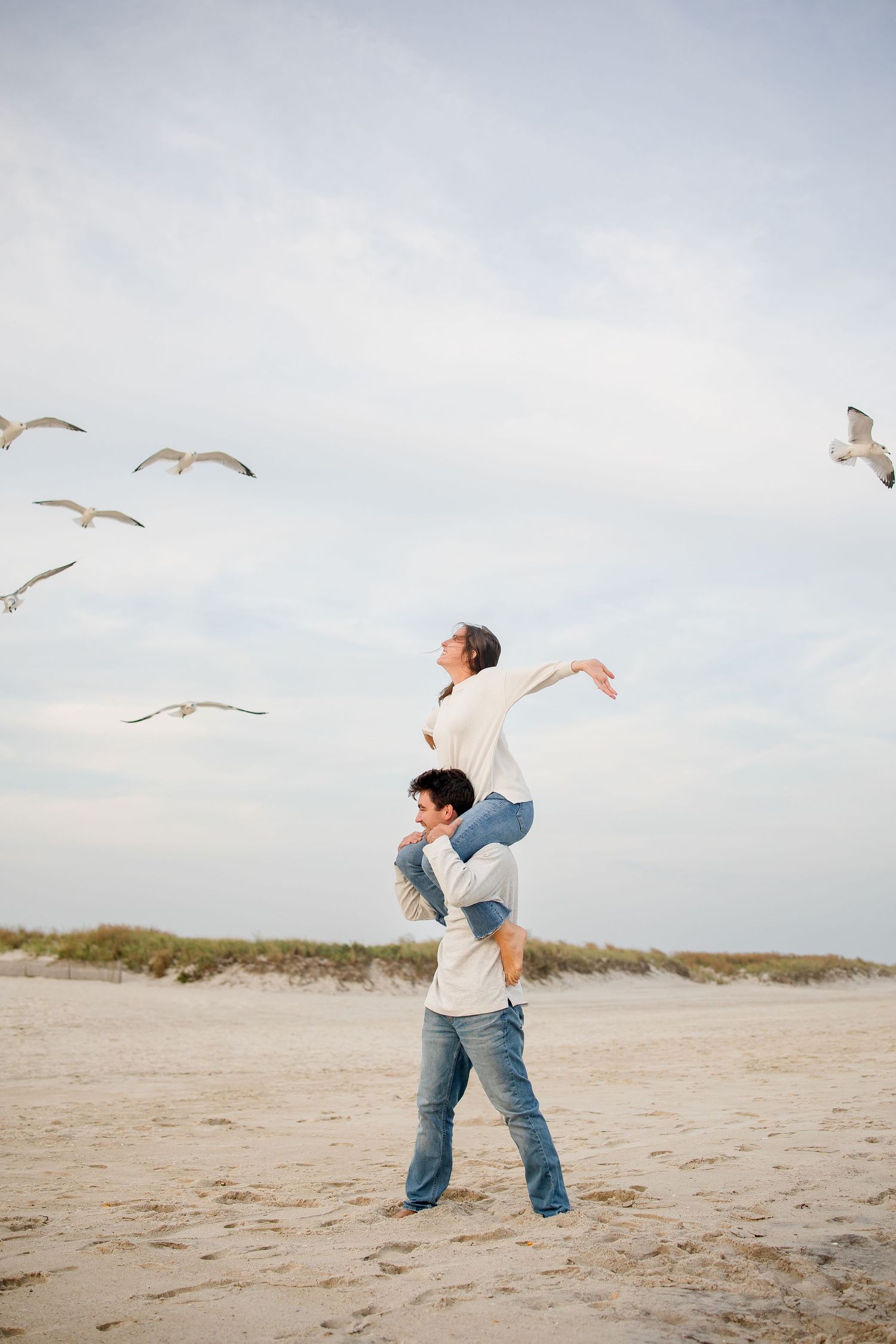 Two people playing on a beach with seagulls flying overhead against a cloudy sky.