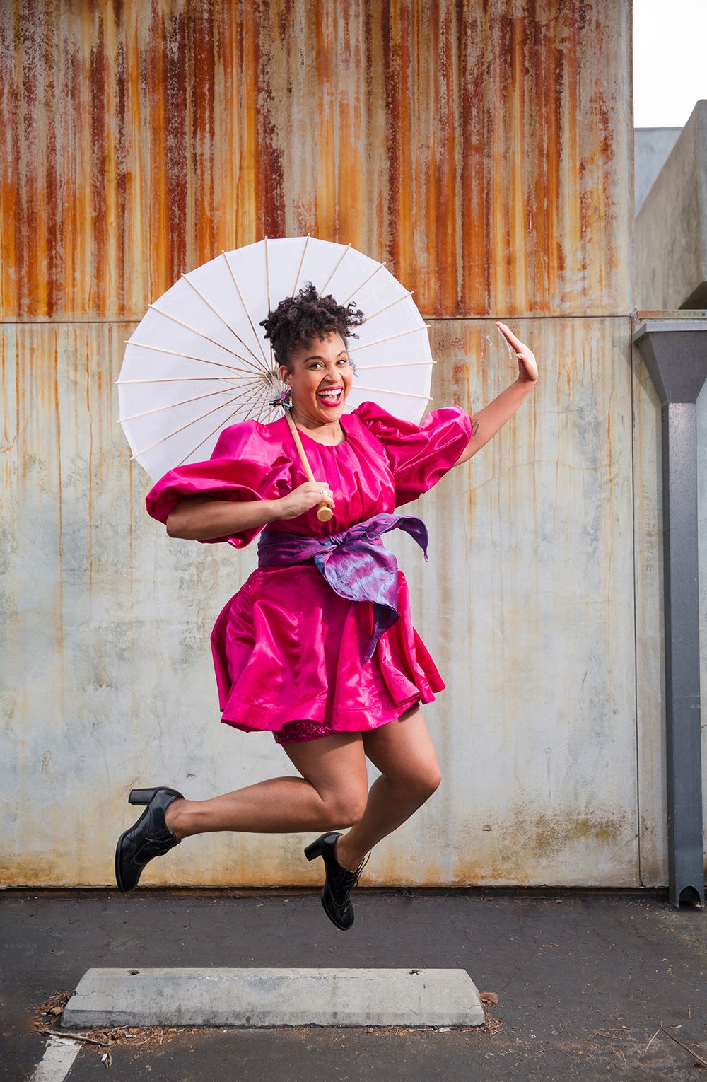 Los Angeles Brand Photographer. A dancer in a vibrant pink dress leaps in the air while holding a parasol against an industrial backdrop.