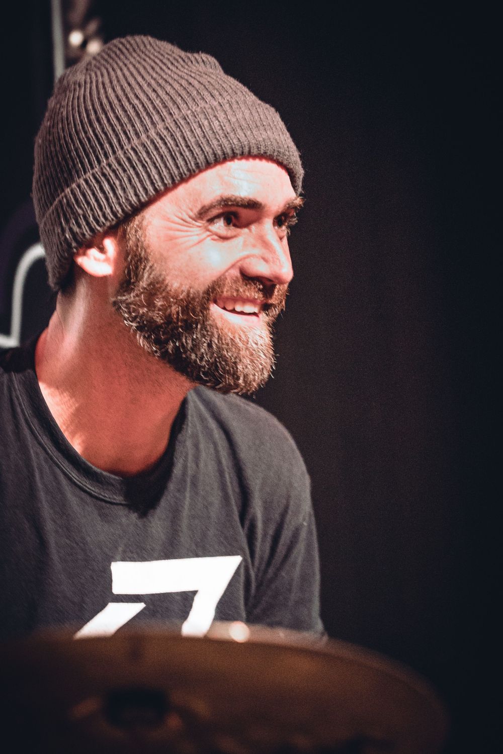 A drummer wearing a brown beanie and black t-shirt smiles while performing behind a drum kit.