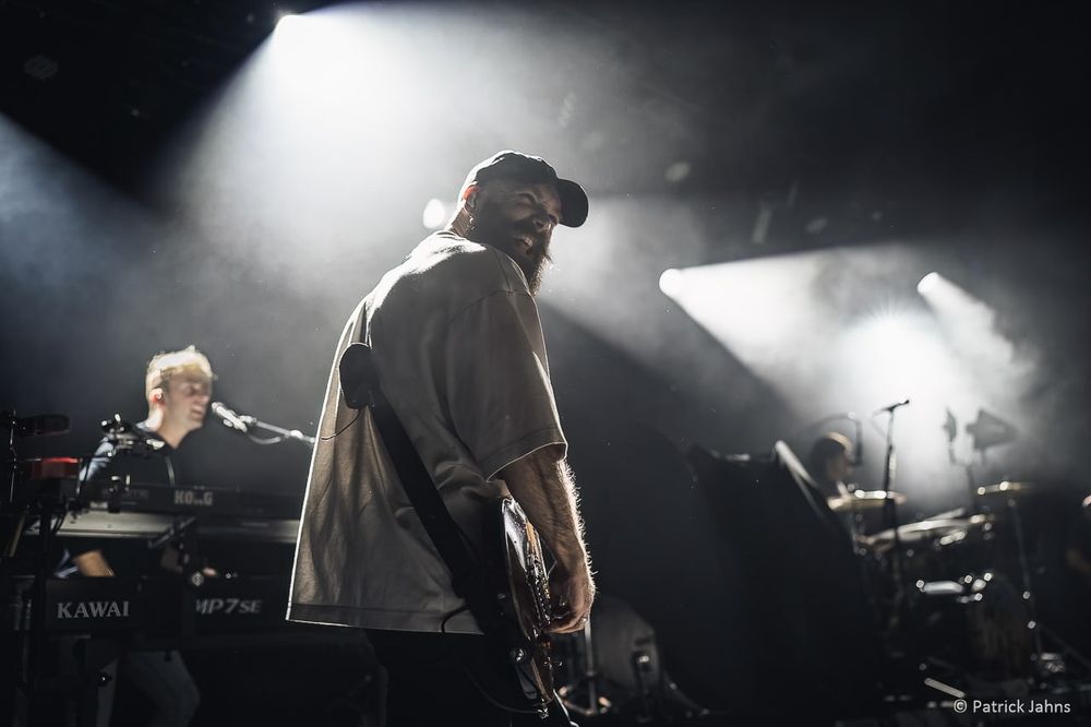 Musicians perform on a dimly lit stage with dramatic backlighting creating silhouettes during a live concert performance.