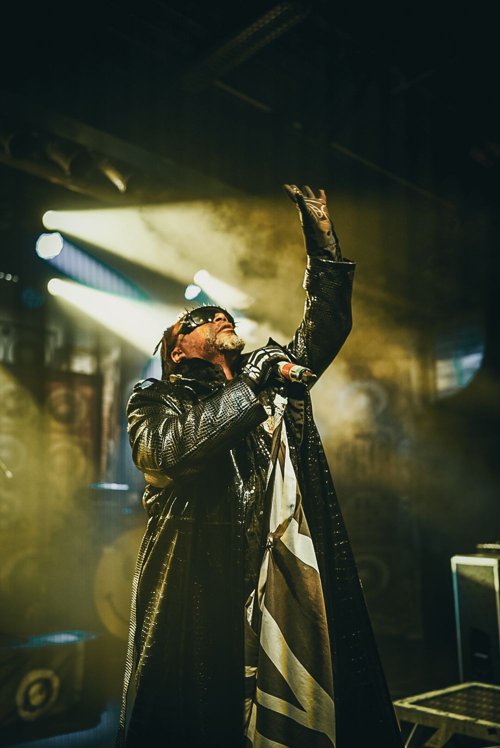 A performer in a leather coat sings passionately on stage with dramatic lighting and fog effects in the background.