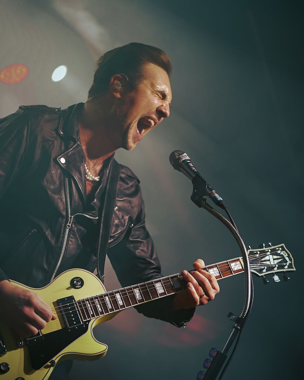 Musician in a leather jacket plays an electric guitar on stage with dramatic lighting and passionate expression.