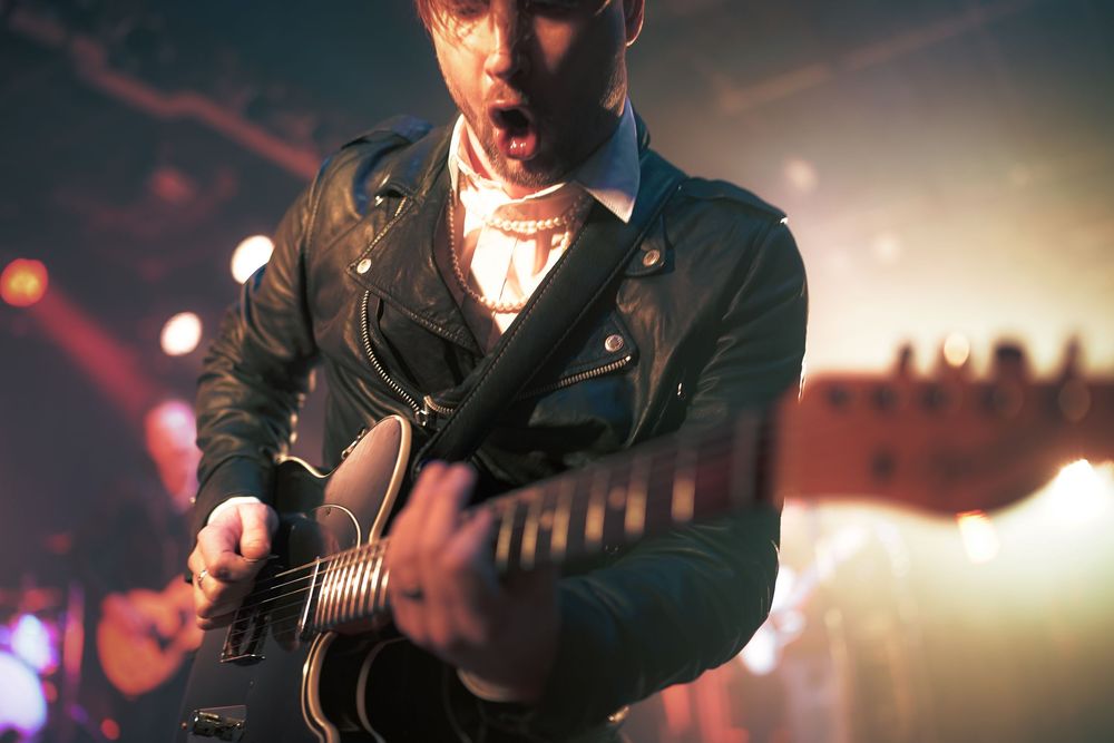 A musician performs on stage wearing a leather jacket while playing an acoustic guitar in dramatic concert lighting.