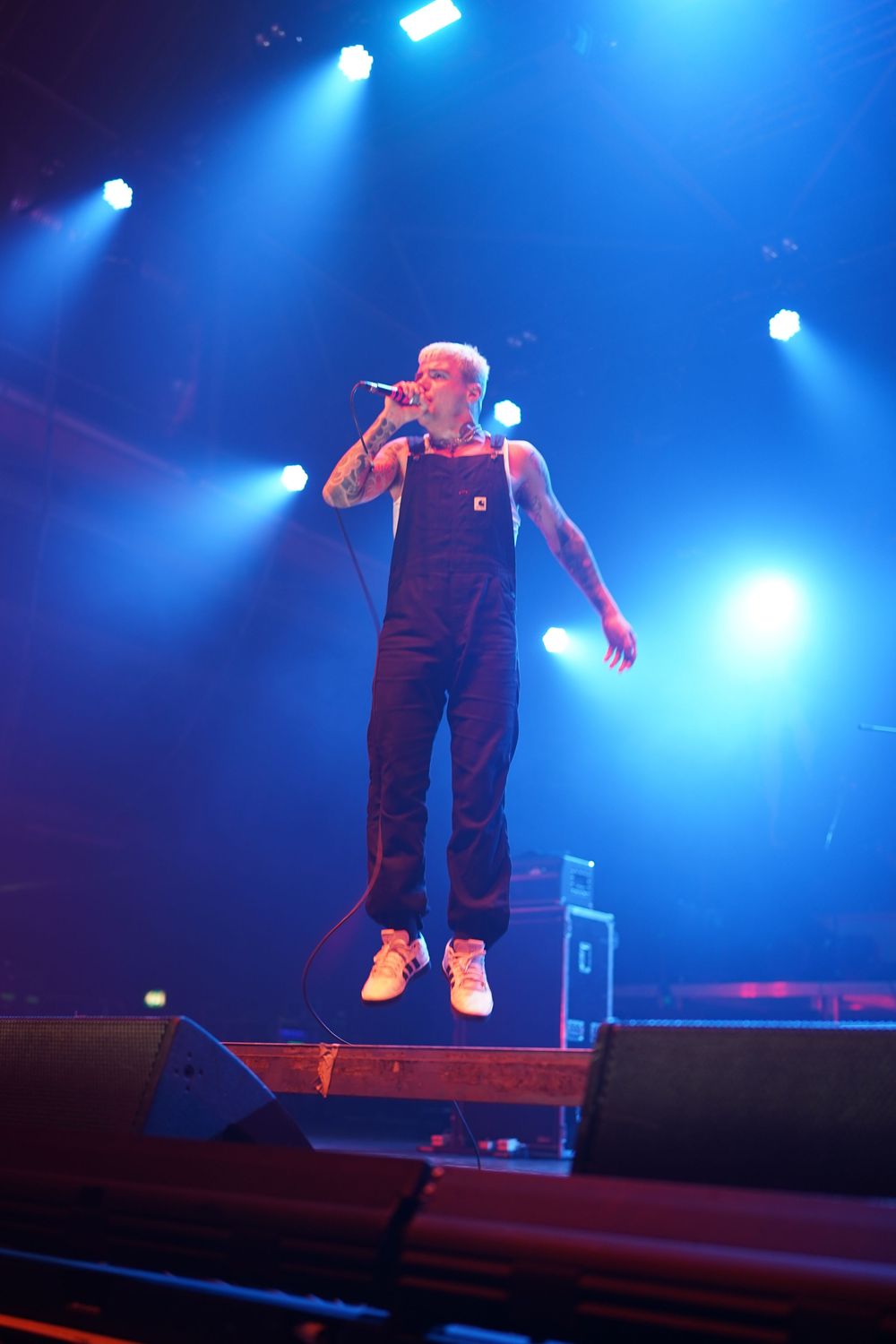 Performer in overalls energetically performs on stage under dramatic blue concert lighting.