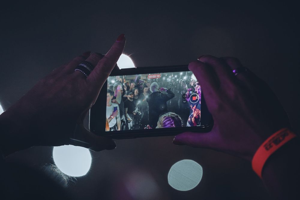 Hands hold up a smartphone recording video at a crowded concert with colorful purple lighting in the background.