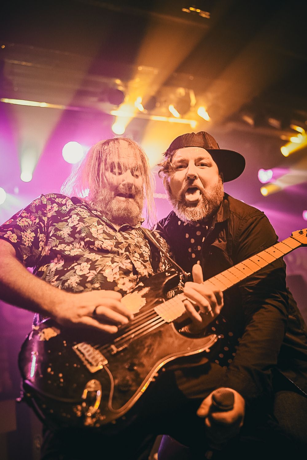 Musicians performing on stage with purple and orange lighting creating a dynamic rock concert atmosphere.
