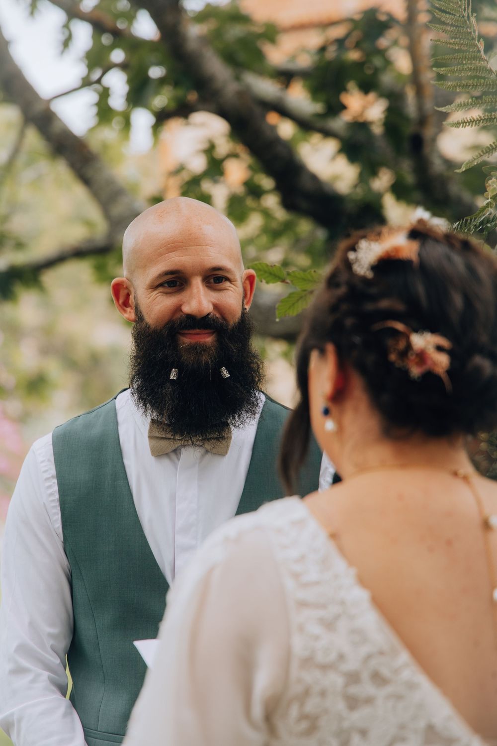 Bald groom with a full beard smiles warmly at his bride during an outdoor wedding ceremony under trees.