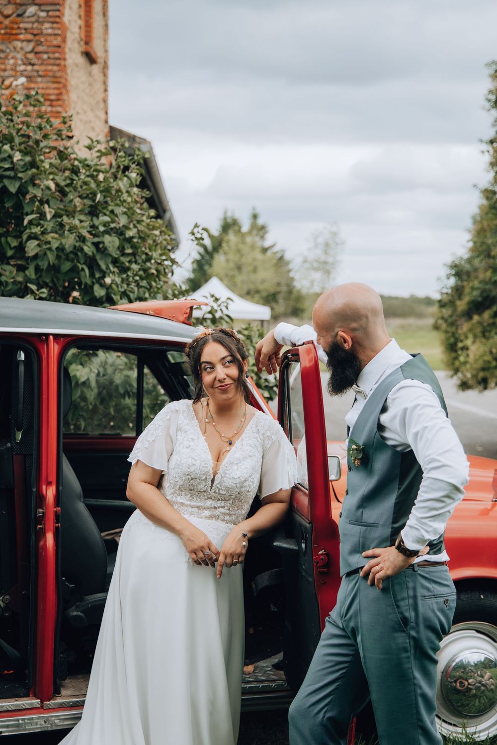 Bride and groom pose beside vintage red car at rustic outdoor wedding venue.