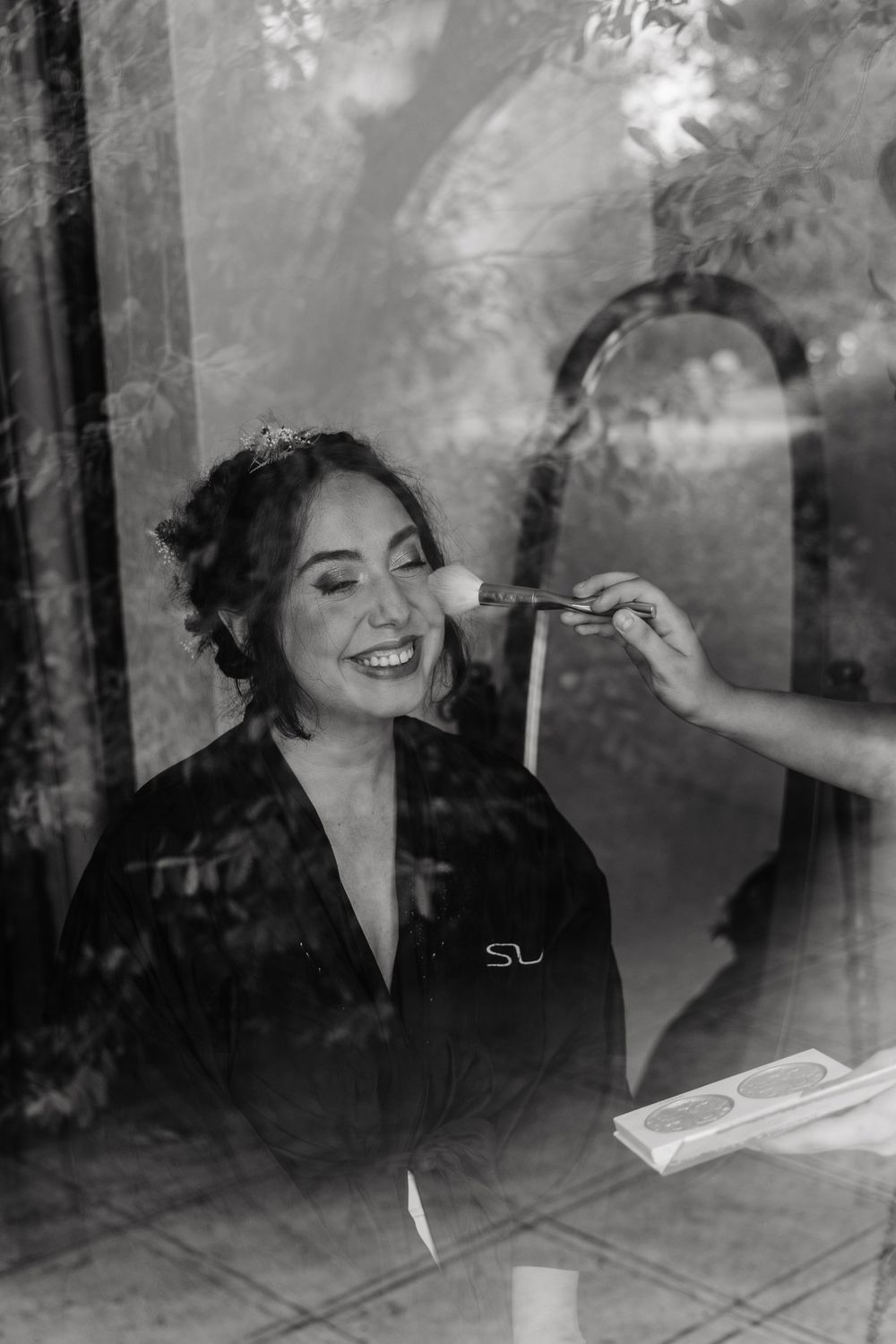 Smiling bride gets makeup applied while wearing black robe in black and white photo.
