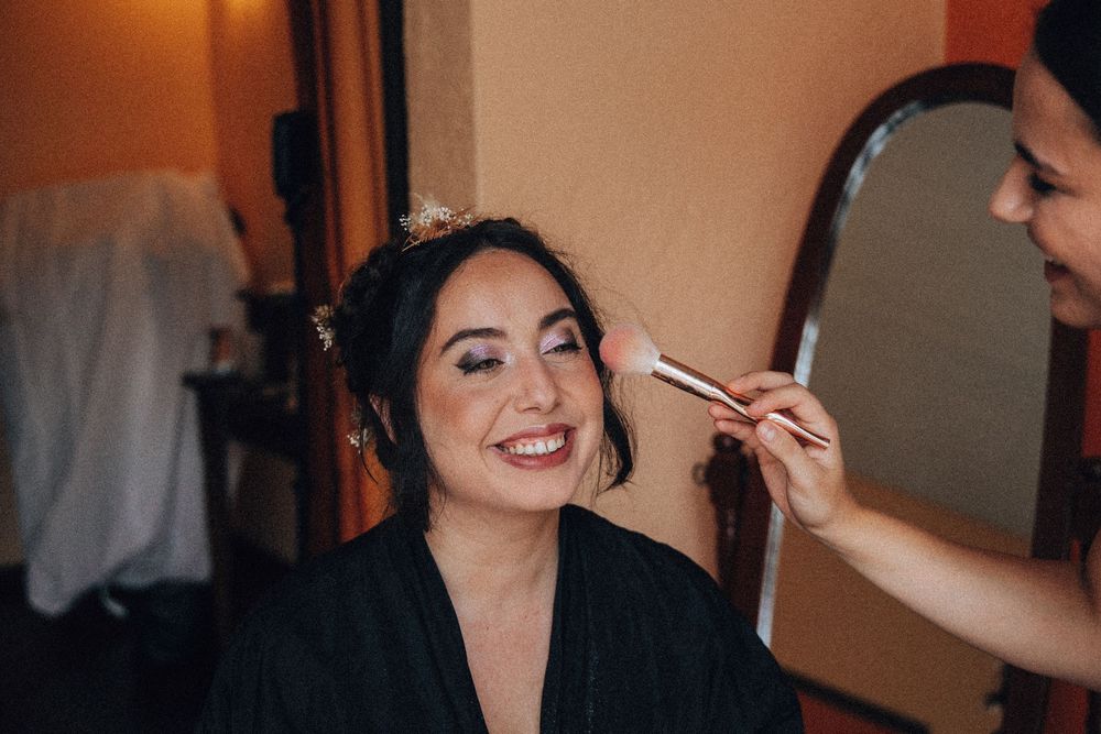 Bride smiles while makeup artist applies brush to her face before wedding ceremony.