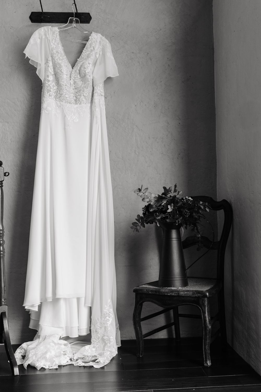 Elegant white wedding dress with lace bodice hanging on wall beside vintage chair in black and white.