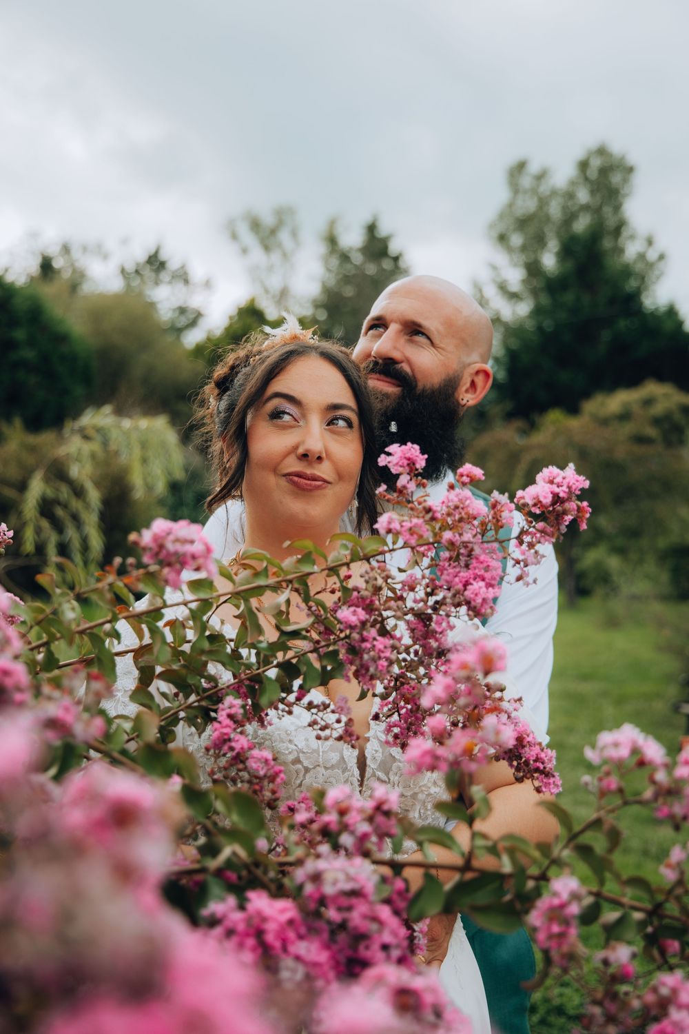 Bride and bearded groom share a romantic moment surrounded by blooming pink flowers in a garden.