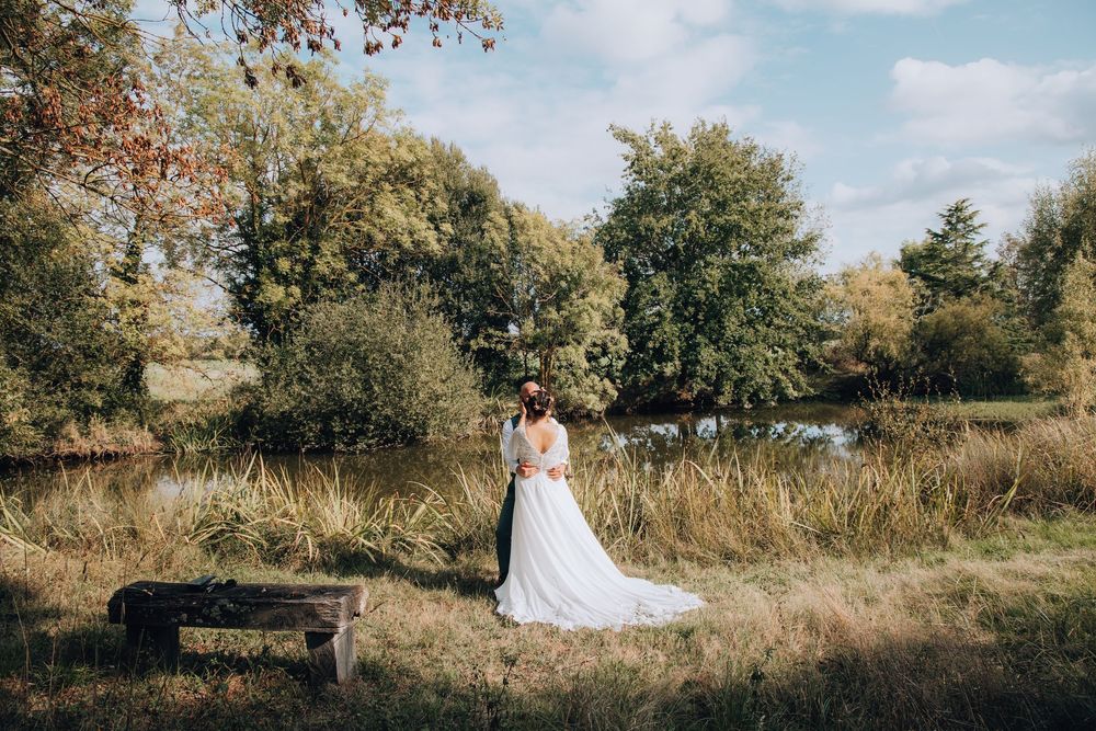 Bride in a flowing white gown stands near a serene pond surrounded by autumn trees on her wedding day.