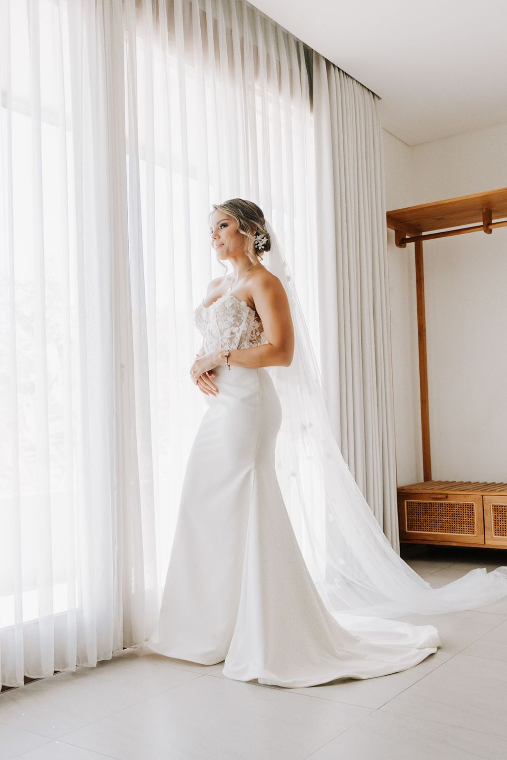 A bride in an elegant white wedding gown with a flowing train stands against sheer white curtains in a bright room.