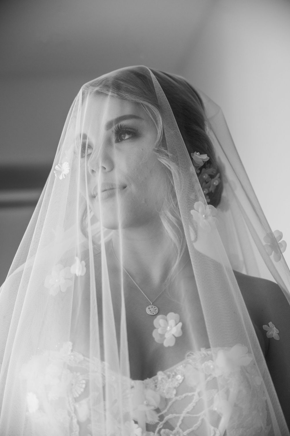 A bride wearing a delicate veil with floral appliques gazes upward in an elegant black and white portrait.