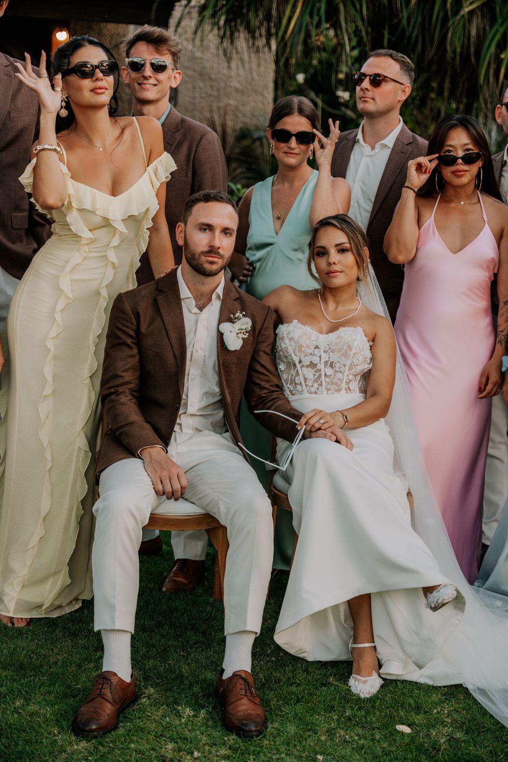 Wedding party members wearing sunglasses pose casually together outdoors in formal attire at sunset.