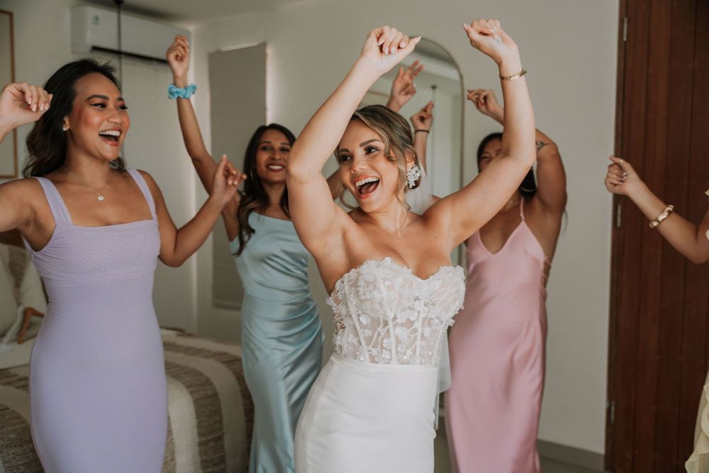 Bridesmaids in pastel dresses dance and celebrate joyfully in a bright indoor venue.