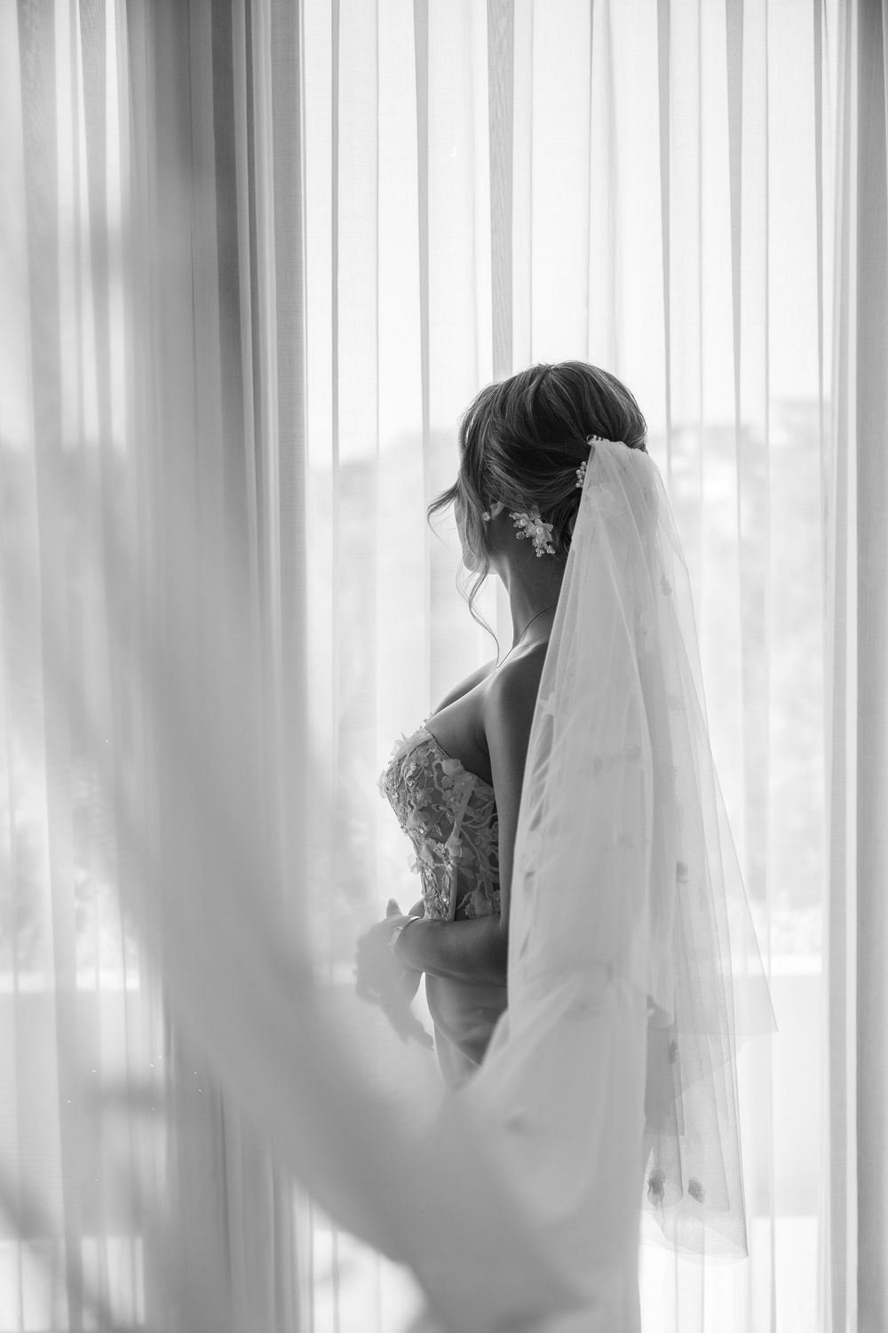 A dreamy black and white photo of a bride in her wedding dress standing by sheer curtained windows.