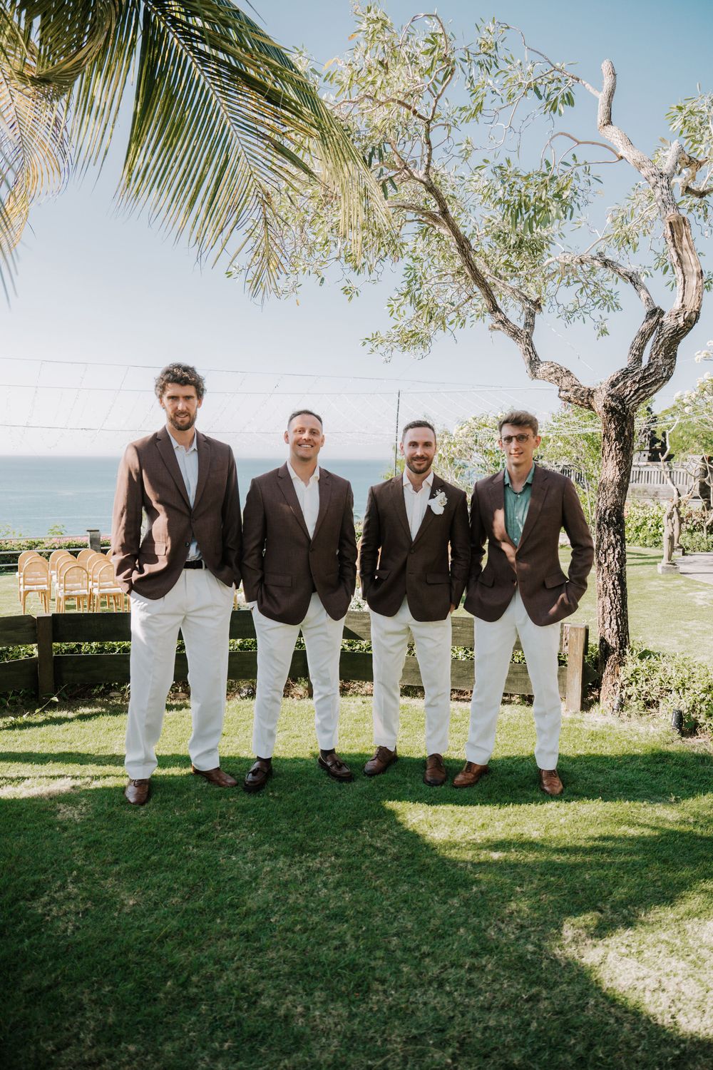 Four people in matching blazers and white pants pose together on a grassy lawn with palm trees and ocean views.