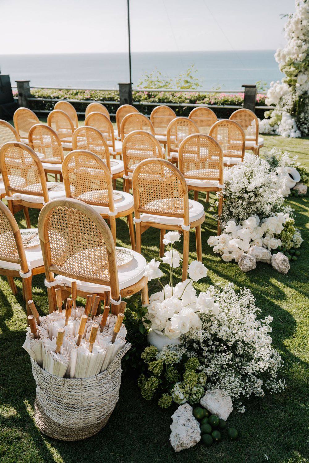Elegant outdoor wedding ceremony setup with cane chairs and white floral arrangements overlooking an ocean view.