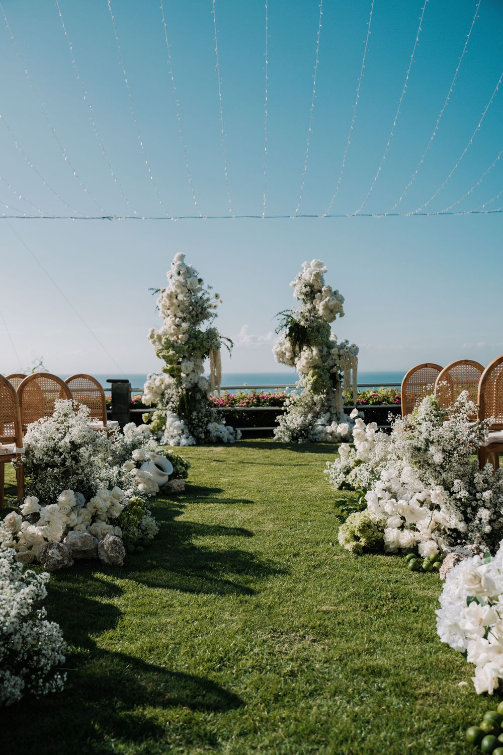 Elegant outdoor wedding ceremony setup with white floral arrangements and rattan chairs on a grassy hilltop against blue skies.