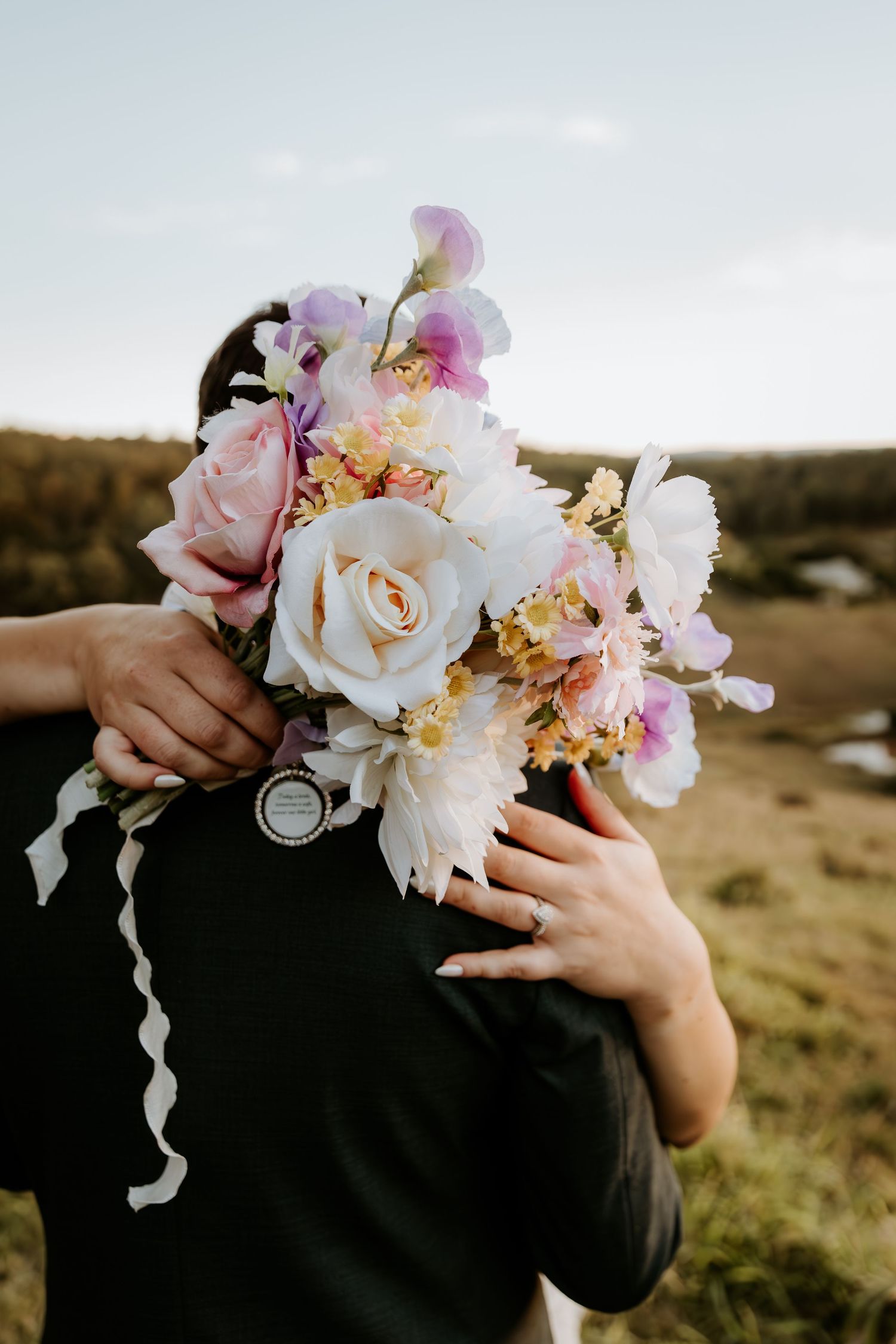 A delicate bouquet of pink and white roses with flowing ribbons held against a moody outdoor landscape at sunset.