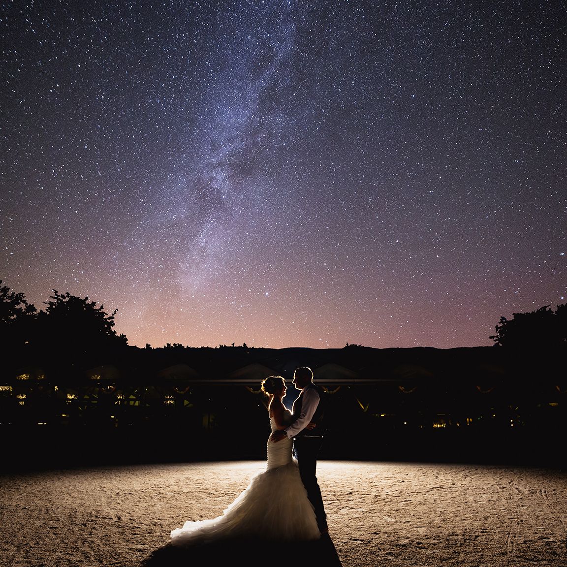 A couple dancing under a starlit sky with the Milky Way visible above and silhouetted trees in the background.
