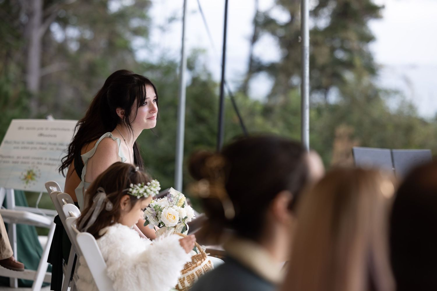A bride in a white wedding dress smiles while holding a bouquet during an outdoor wedding ceremony.