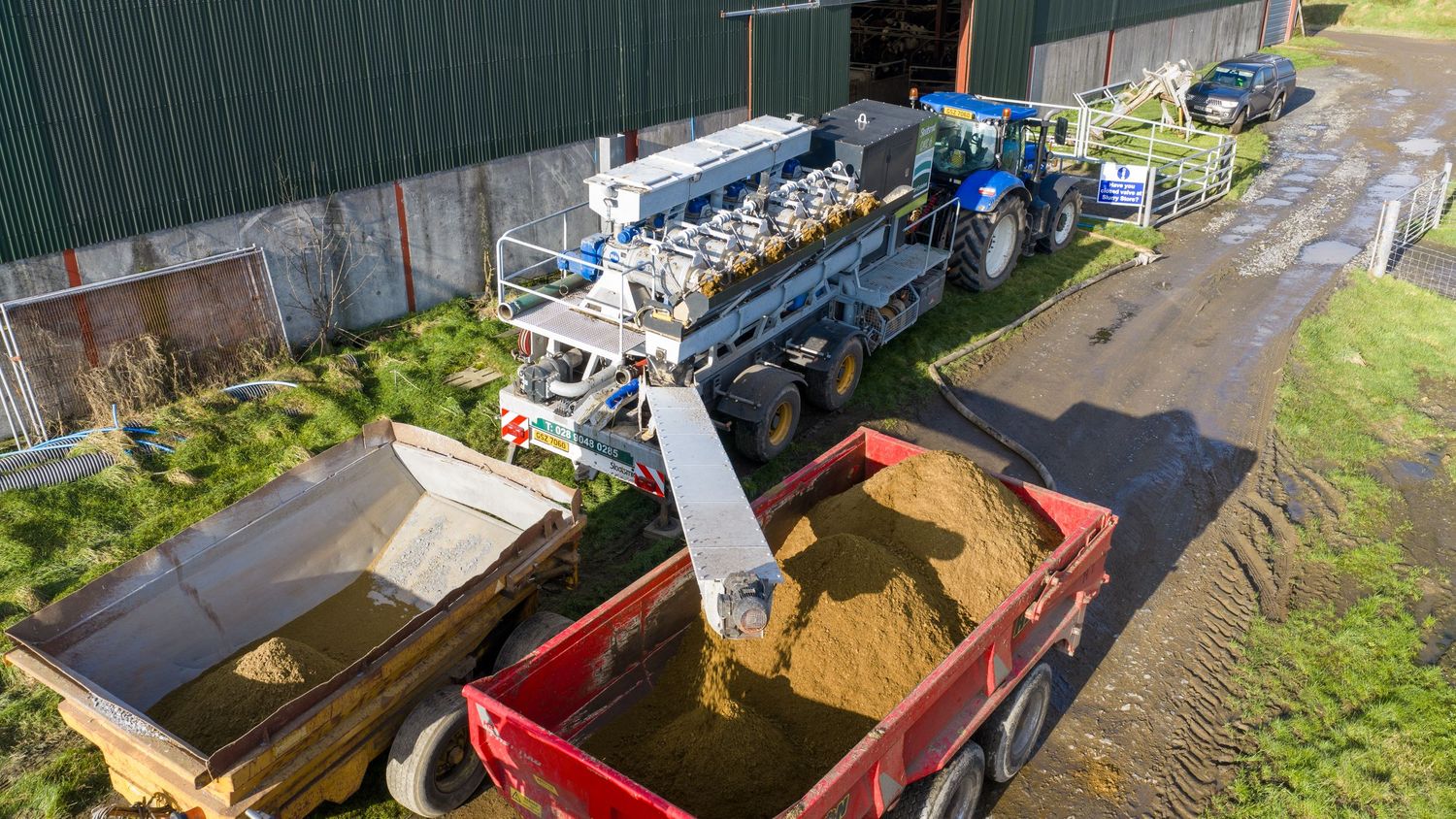 Grain being transferred from a large blue machine into a red truck trailer at an agricultural facility.