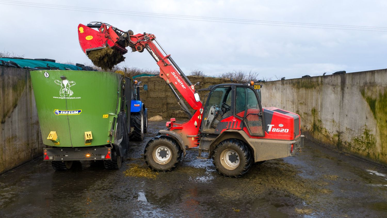 A red telescopic handler machine loads green agricultural trailers on a farm during wet weather conditions.