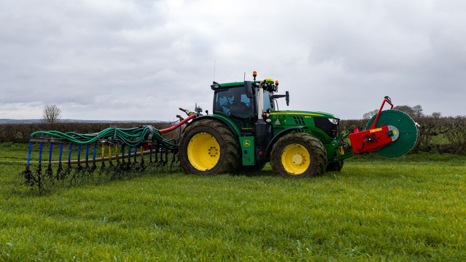 Green John Deere tractor pulling farm equipment across a lush agricultural field under cloudy skies.