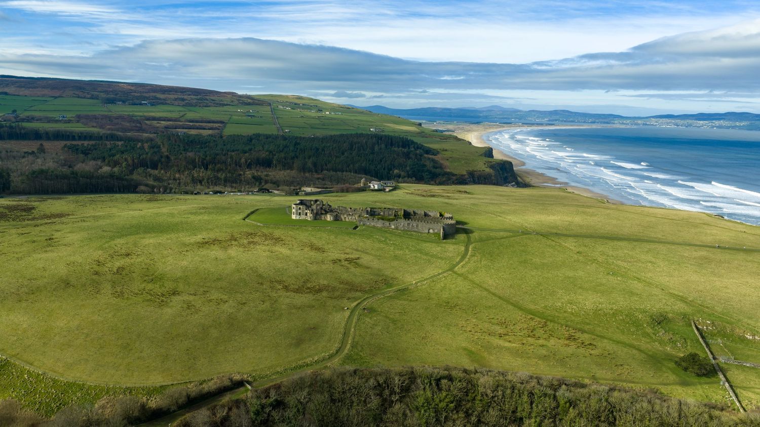 Aerial view of green coastal cliffs with scattered buildings along a dramatic beach shoreline in Scotland.