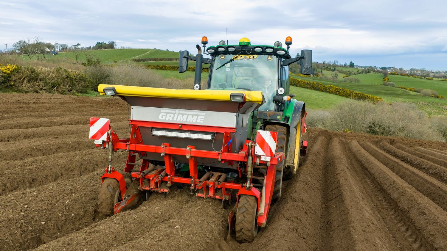 Panoramic view of agricultural machinery planting crops in fertile farmland with rolling hills in the background.