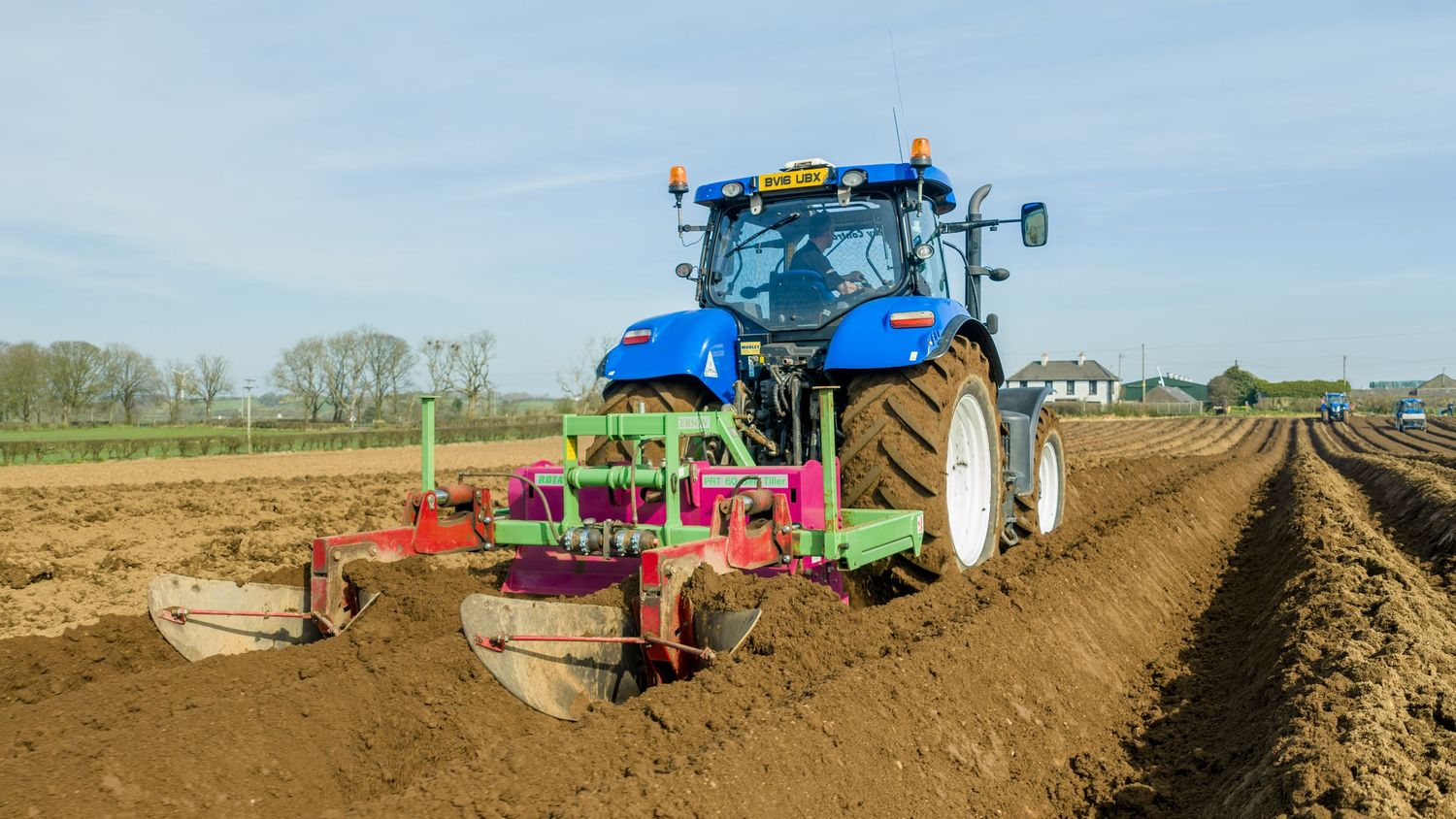 Tractor pulling agricultural equipment plows and tills brown soil in a rural farm field on a sunny day.