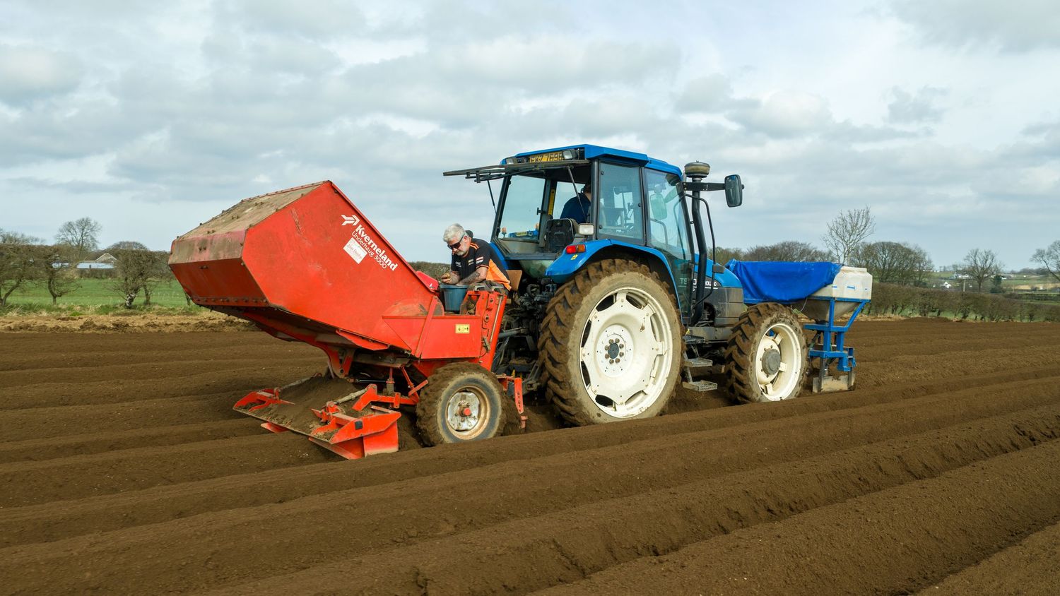 Blue tractor with red planting attachment working in freshly tilled brown soil under cloudy skies.