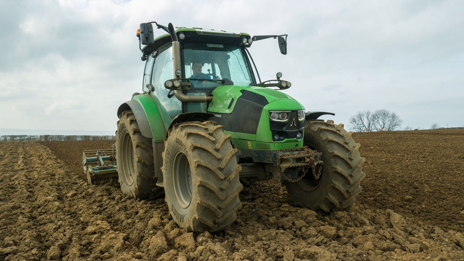 Green John Deere tractor plowing a field of brown soil against an overcast sky.
