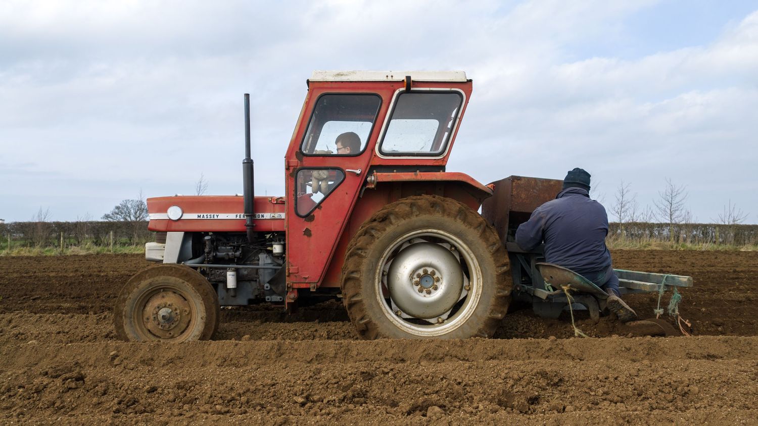 A red tractor plows through brown soil in an agricultural field preparing the ground for planting season.