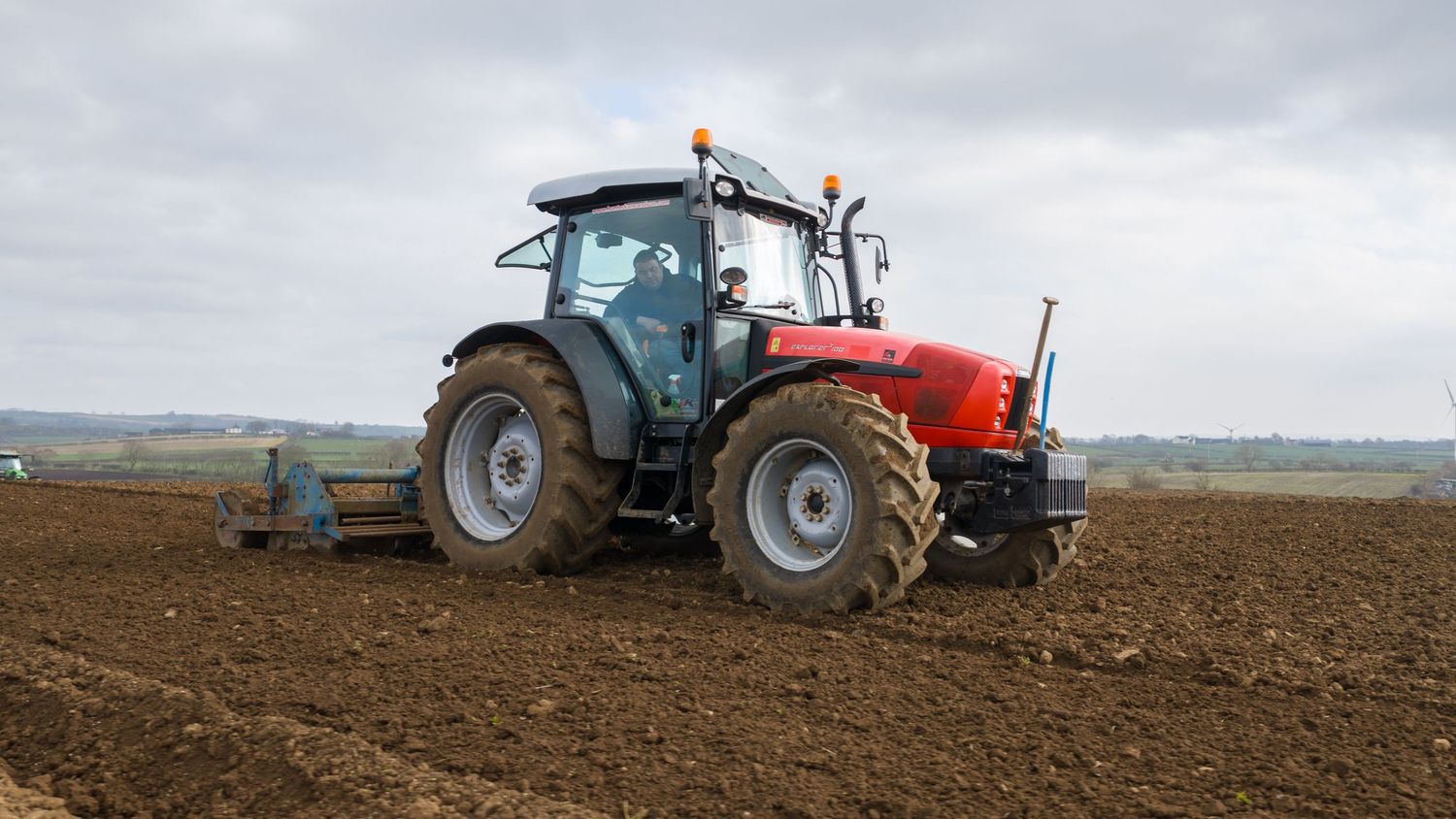 A panoramic view of tractors plowing agricultural fields under an overcast sky during farming season.