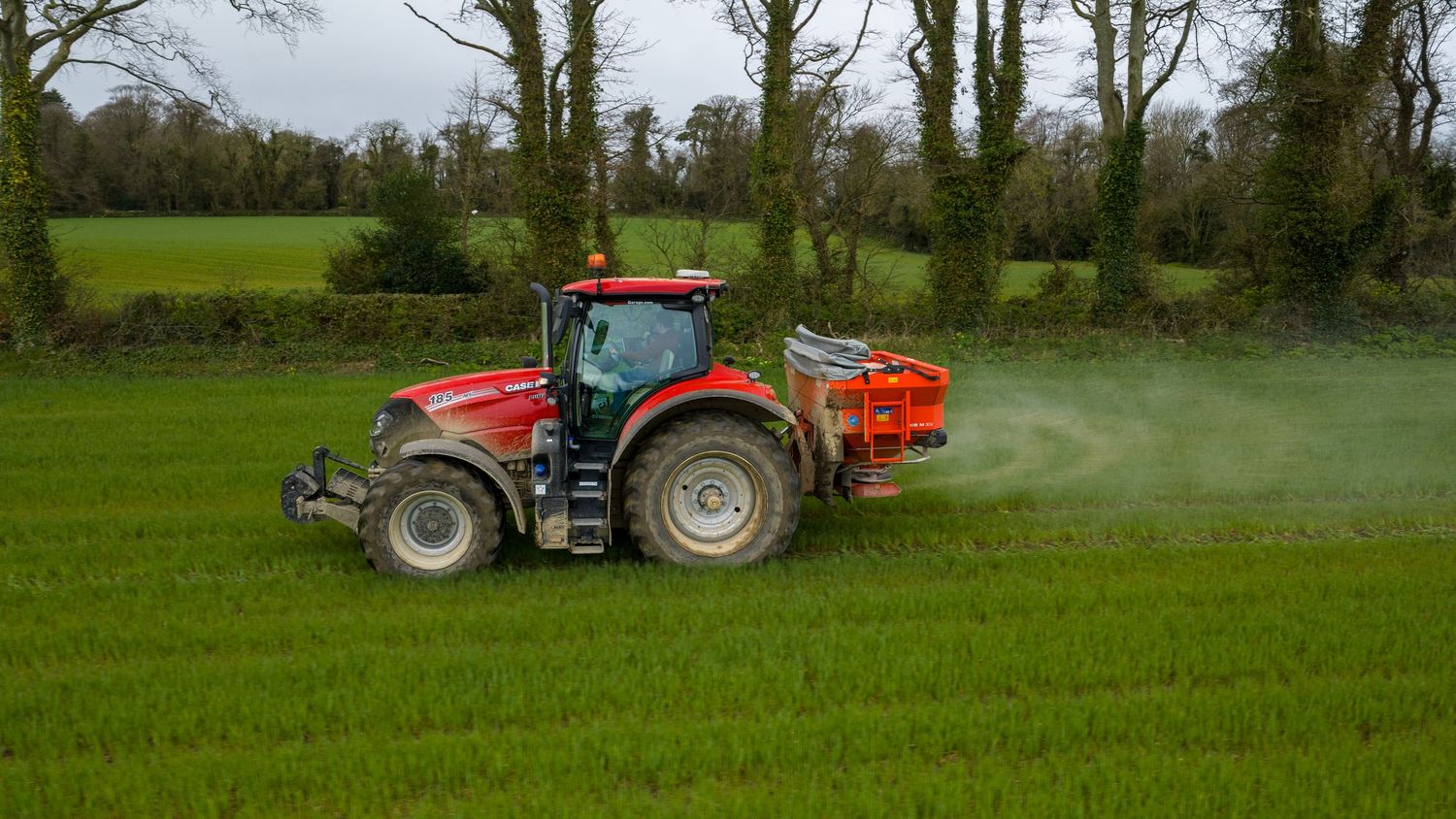 Red tractor spraying fertilizer on green agricultural field while moving across farmland in spring.