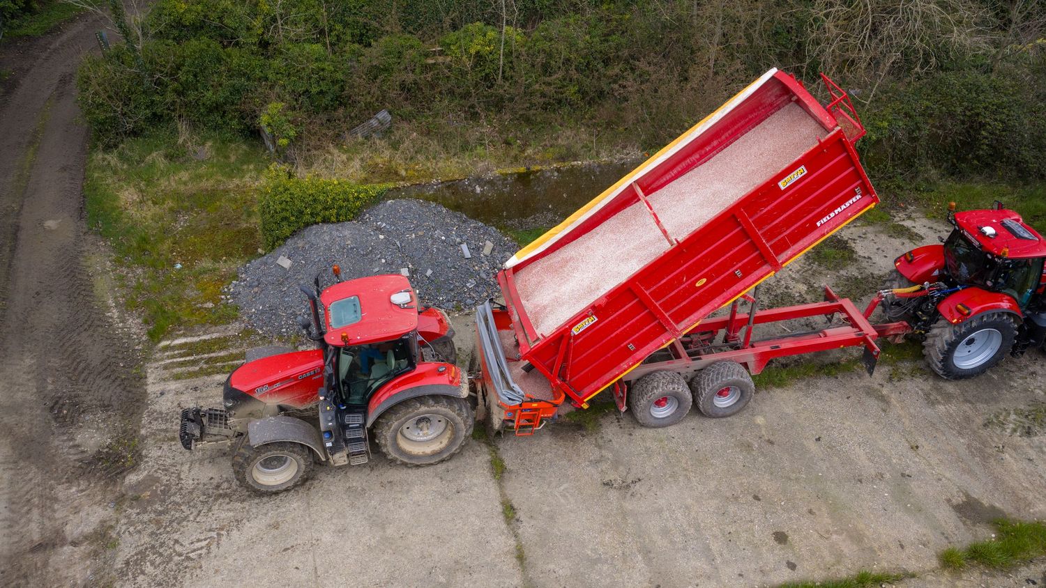 Aerial view of a red tractor with trailer parked on a dirt path next to green forest vegetation.