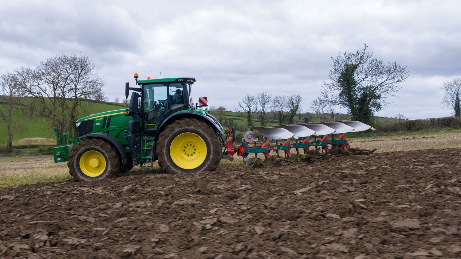 Green John Deere tractor pulling a large plow through brown soil in a rural field under cloudy skies.