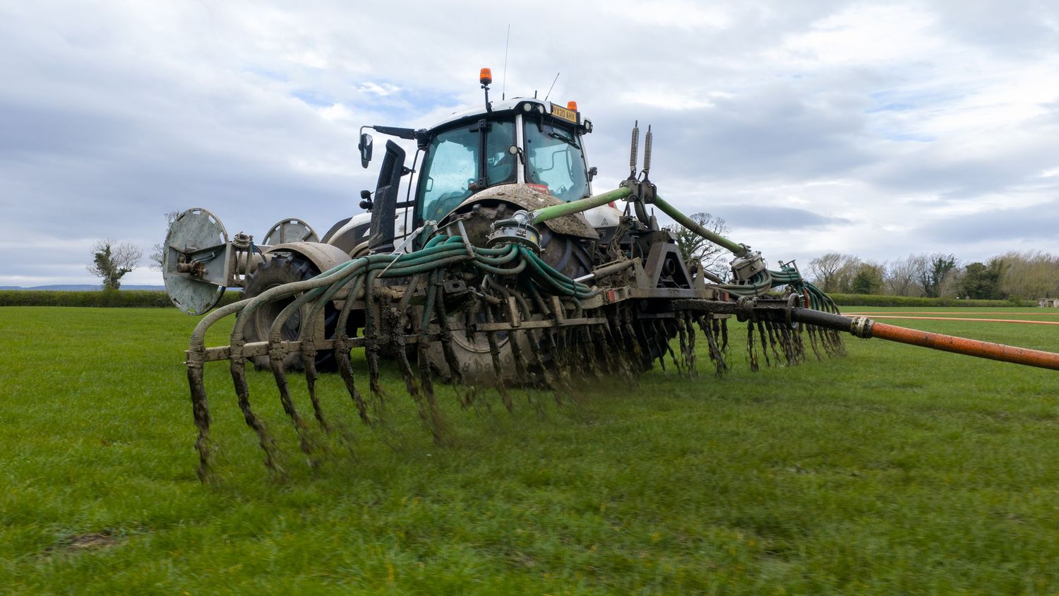 Green tractor with wide cultivator attachment working in a lush agricultural field on a cloudy day.