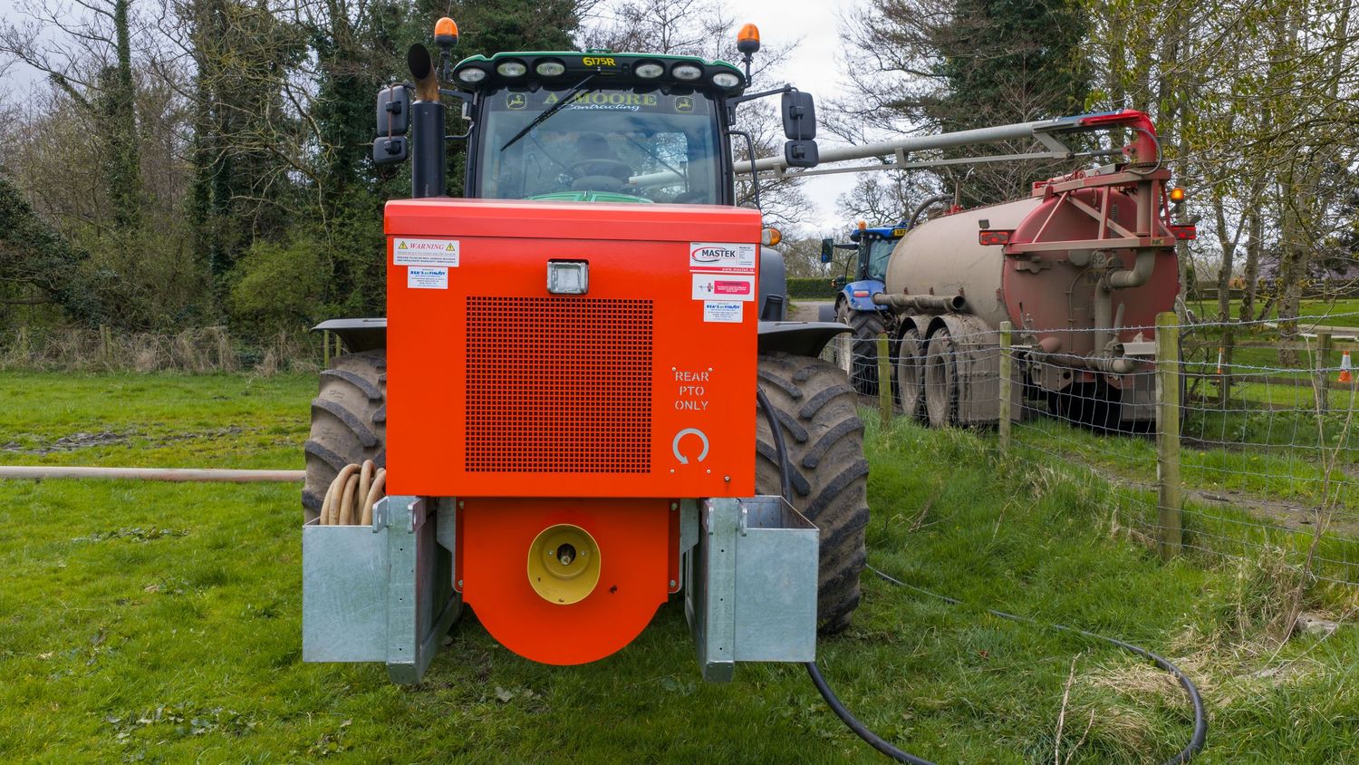 Red farm tractor viewed from rear in green field, connected to agricultural equipment on a cloudy day.