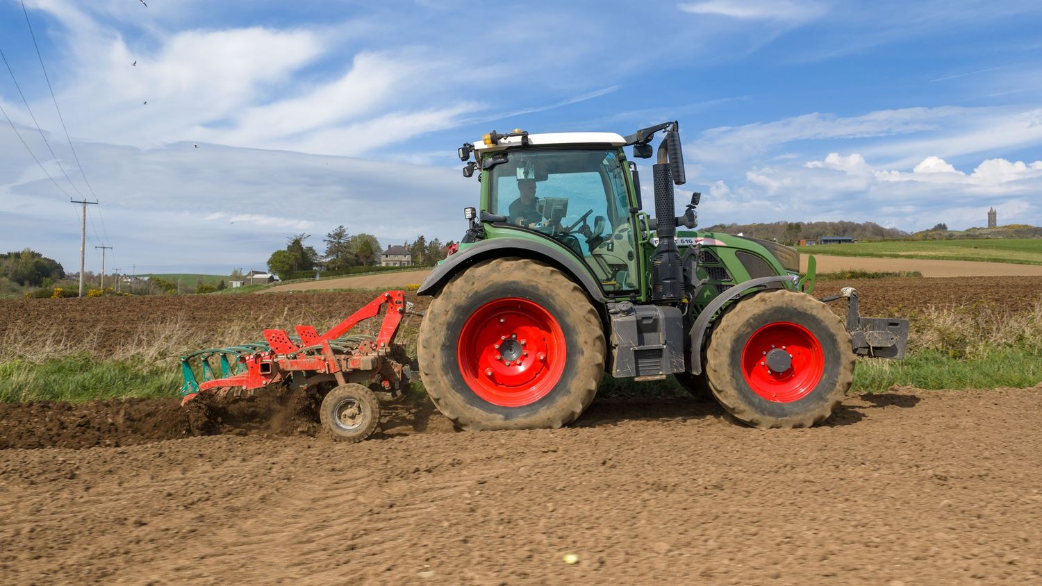 Green tractor with red wheels plowing a field on a sunny day with blue skies and scattered clouds in the background.