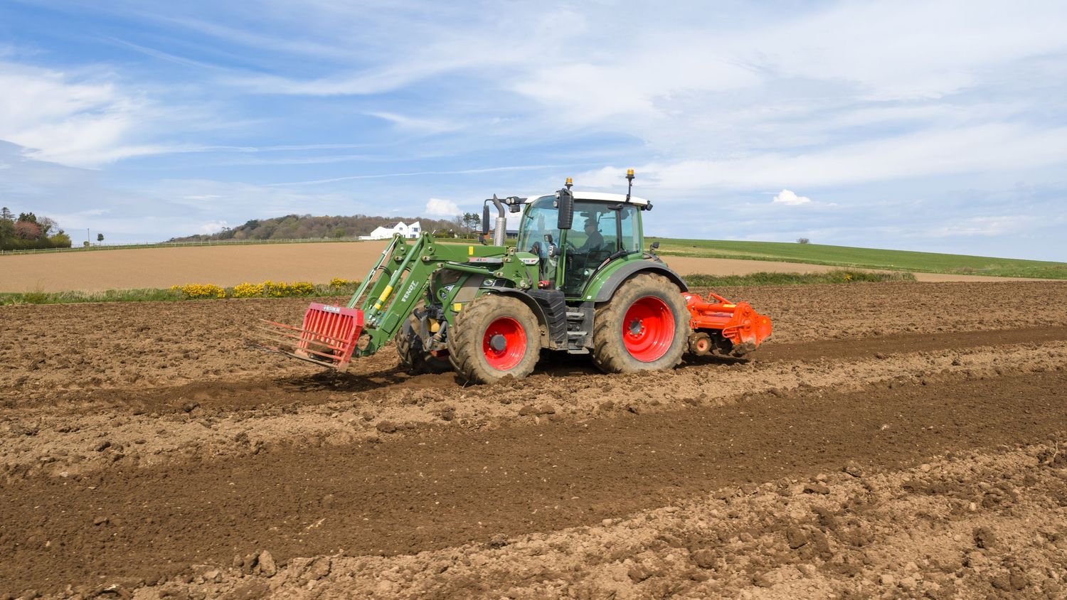 Green Fendt tractor plowing a large agricultural field under a partly cloudy blue sky on a sunny day.