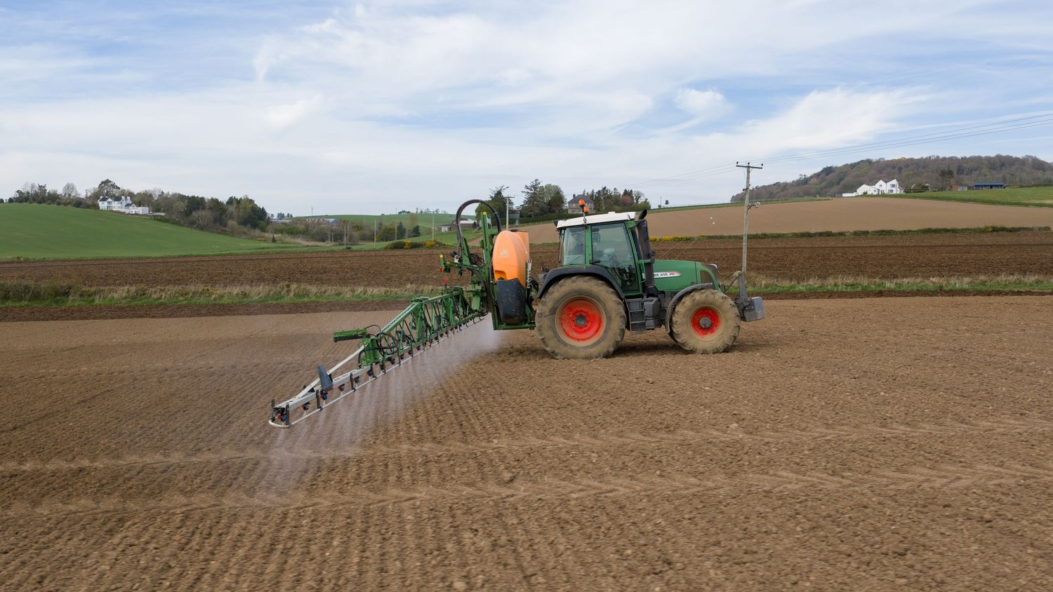Green tractor spraying pesticides on a large agricultural field under a partly cloudy sky in a rural landscape.