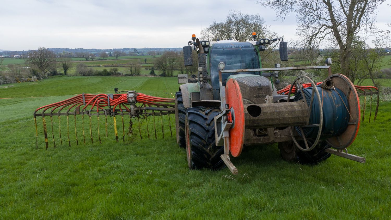 A blue tractor with orange hose reel and drag hose equipment spreading slurry on a green agricultural field.