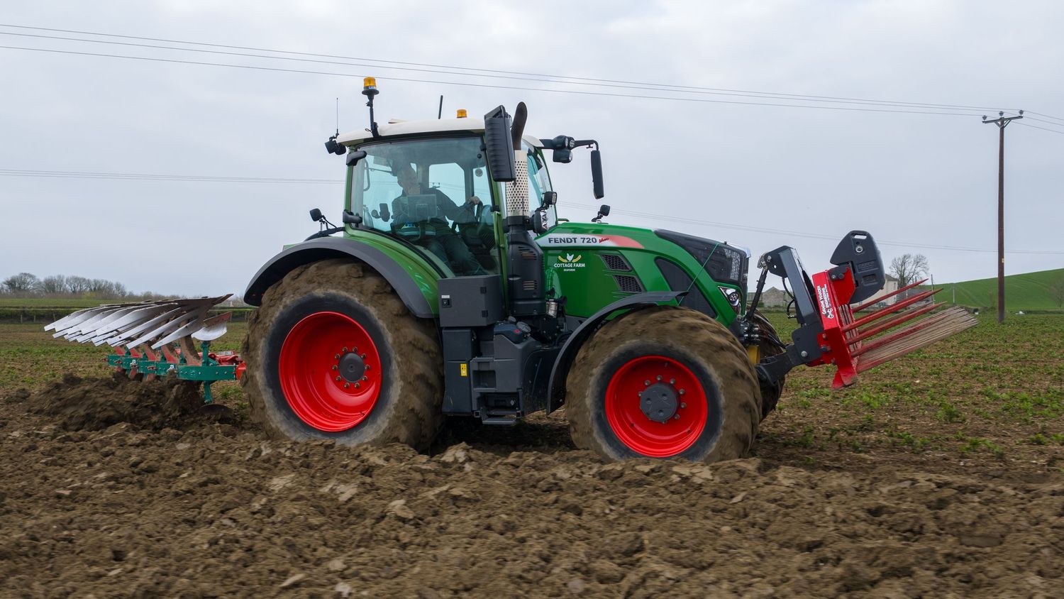 Green Fendt tractor with red wheels plowing a field, attached with a front cultivator on a cloudy day.